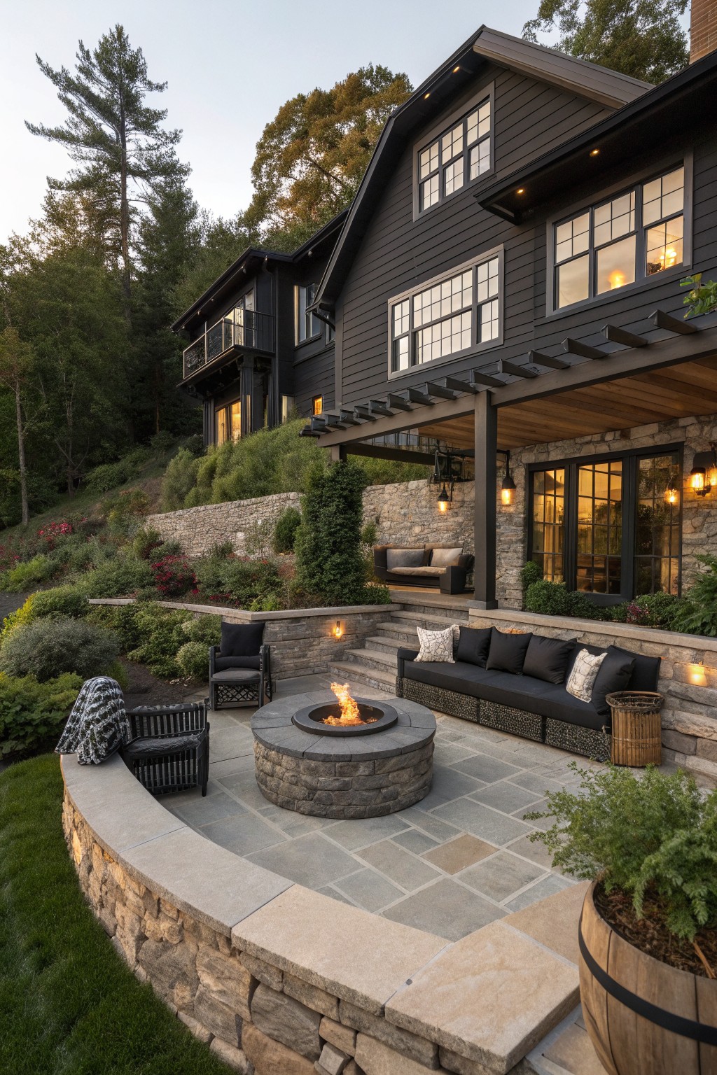 A dark house with stone accents has a stone patio centered around a round fire pit, with built-in and freestanding seating arranged around it.