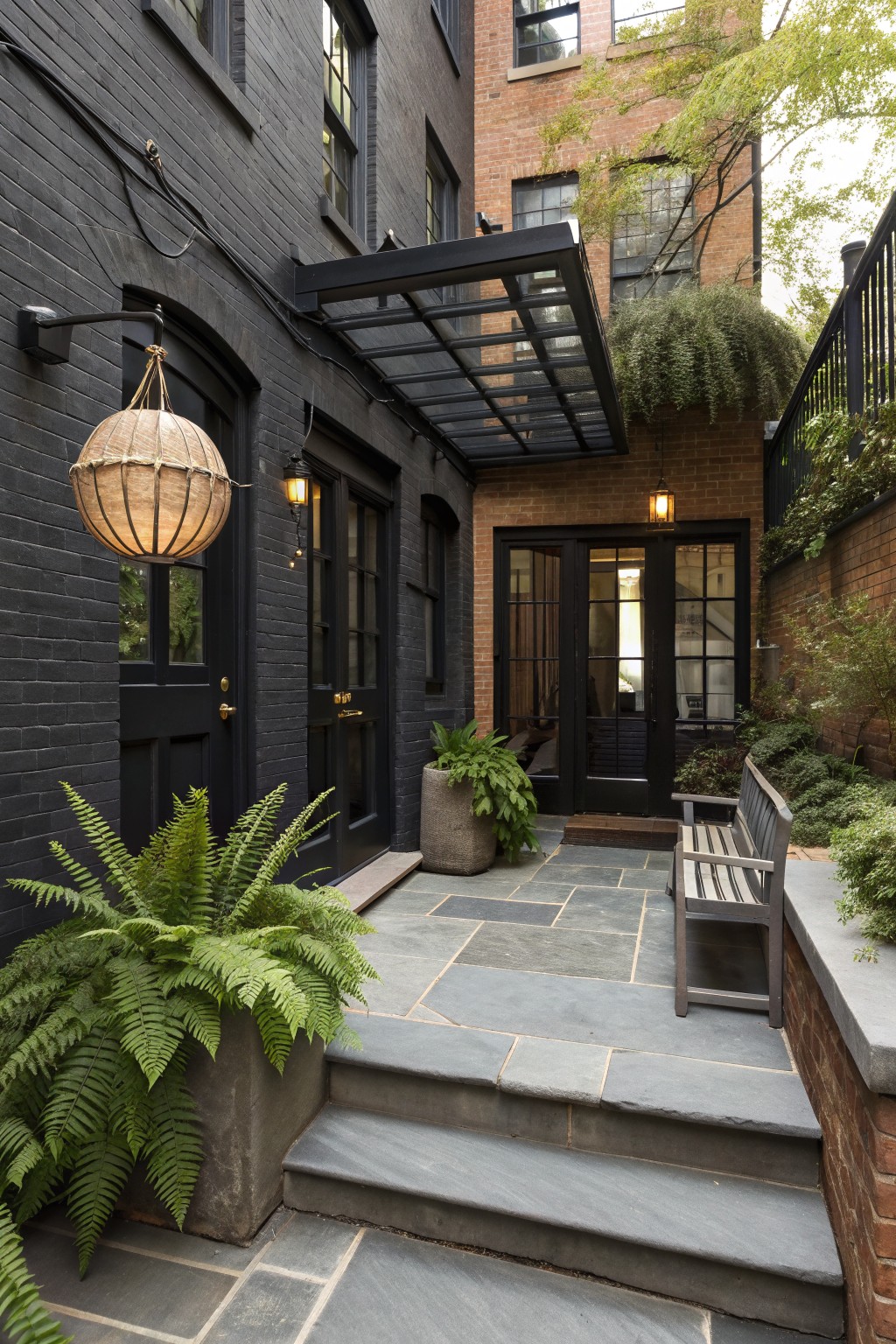 Slate courtyard with black doors, ferns, bench, lanterns, and glass awning on brick walls