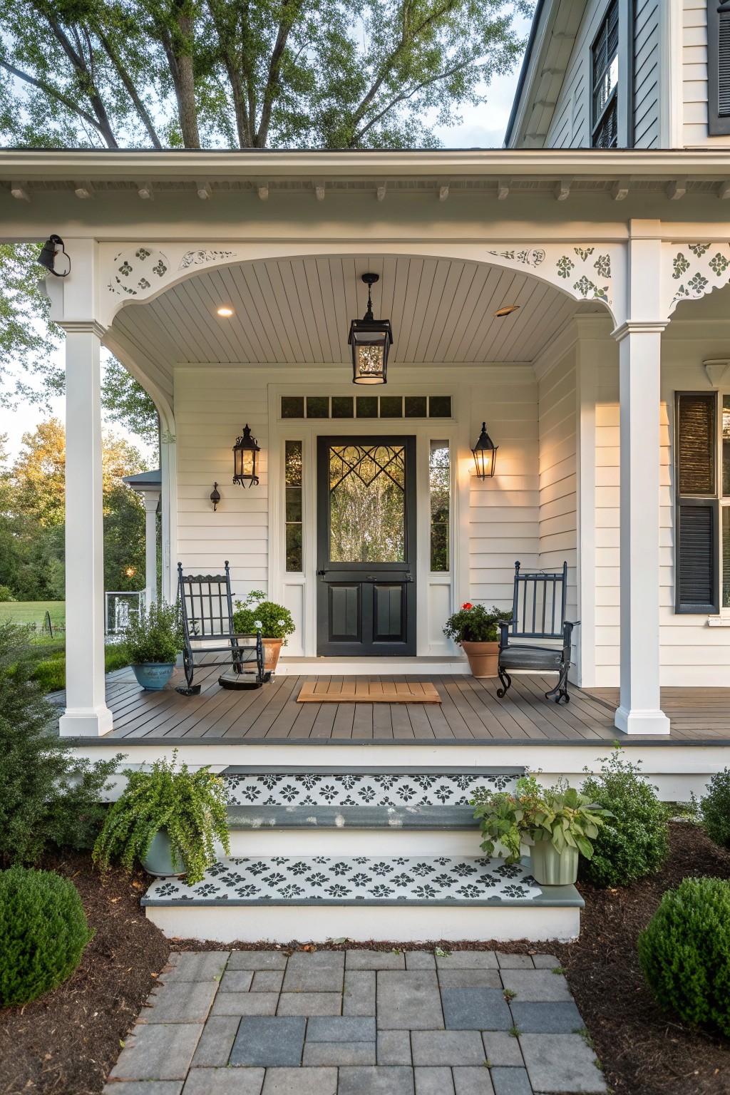 A white Victorian farmhouse front porch with a dark centered door, two matching black rocking chairs, potted plants on the deck, and patterned tile on the steps.