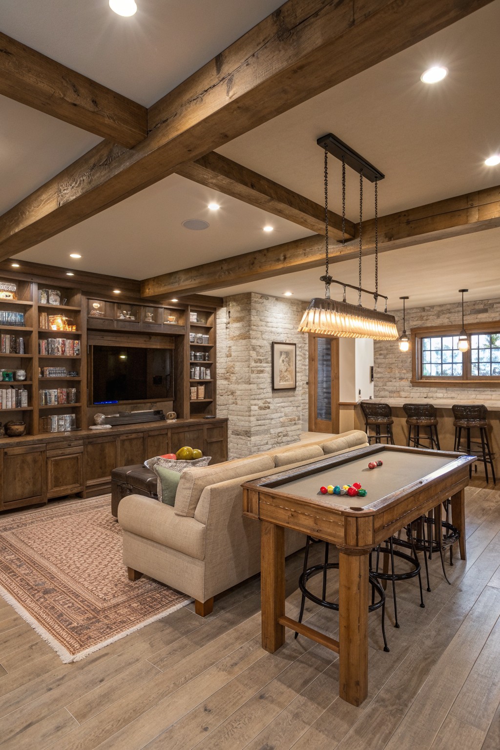 A basement rec room featuring a wooden pool table in the foreground, a beige sofa, built-in wood cabinetry with a television, a stone accent wall, and a bar area with stools under exposed ceiling beams.