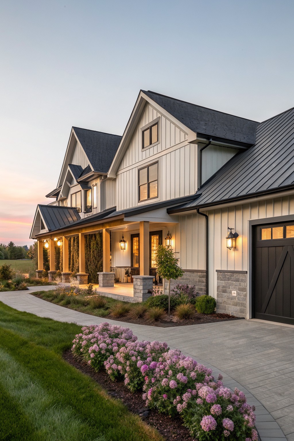 A modern farmhouse exterior featuring white vertical siding, a dark metal roof, stone accents on the lower walls, a covered porch with columns, and a landscaped front with pink flowers along the driveway.