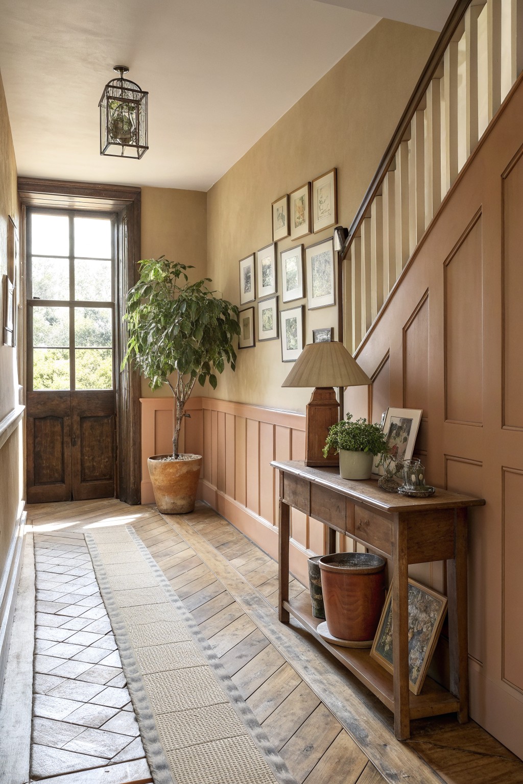 Terracotta hallway with wooden console table, staircase, potted plants, and framed art gallery.