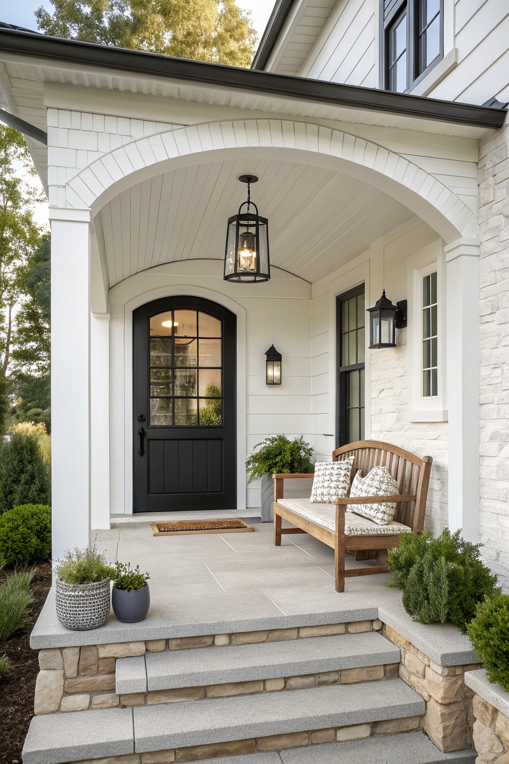 Front view of a white farmhouse porch with a large arched ceiling, black door, wooden bench with cushions, and stone steps.