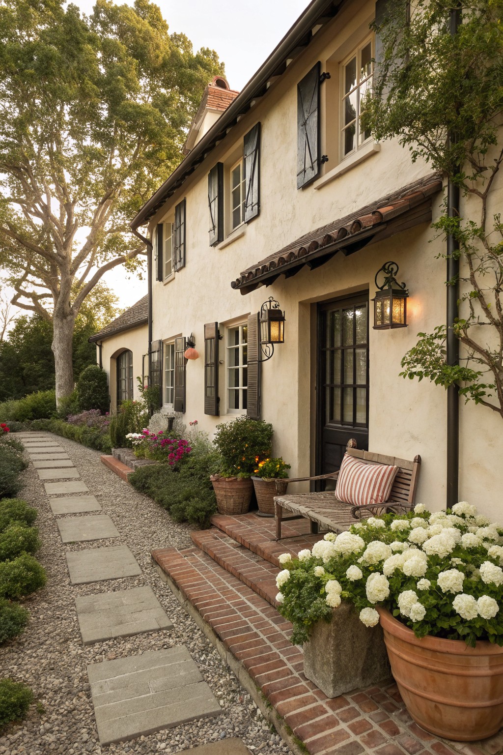 A light stucco house exterior with dark shutters and trim around the windows and door, brick steps, potted plants, and a stone pathway leading to the entrance.