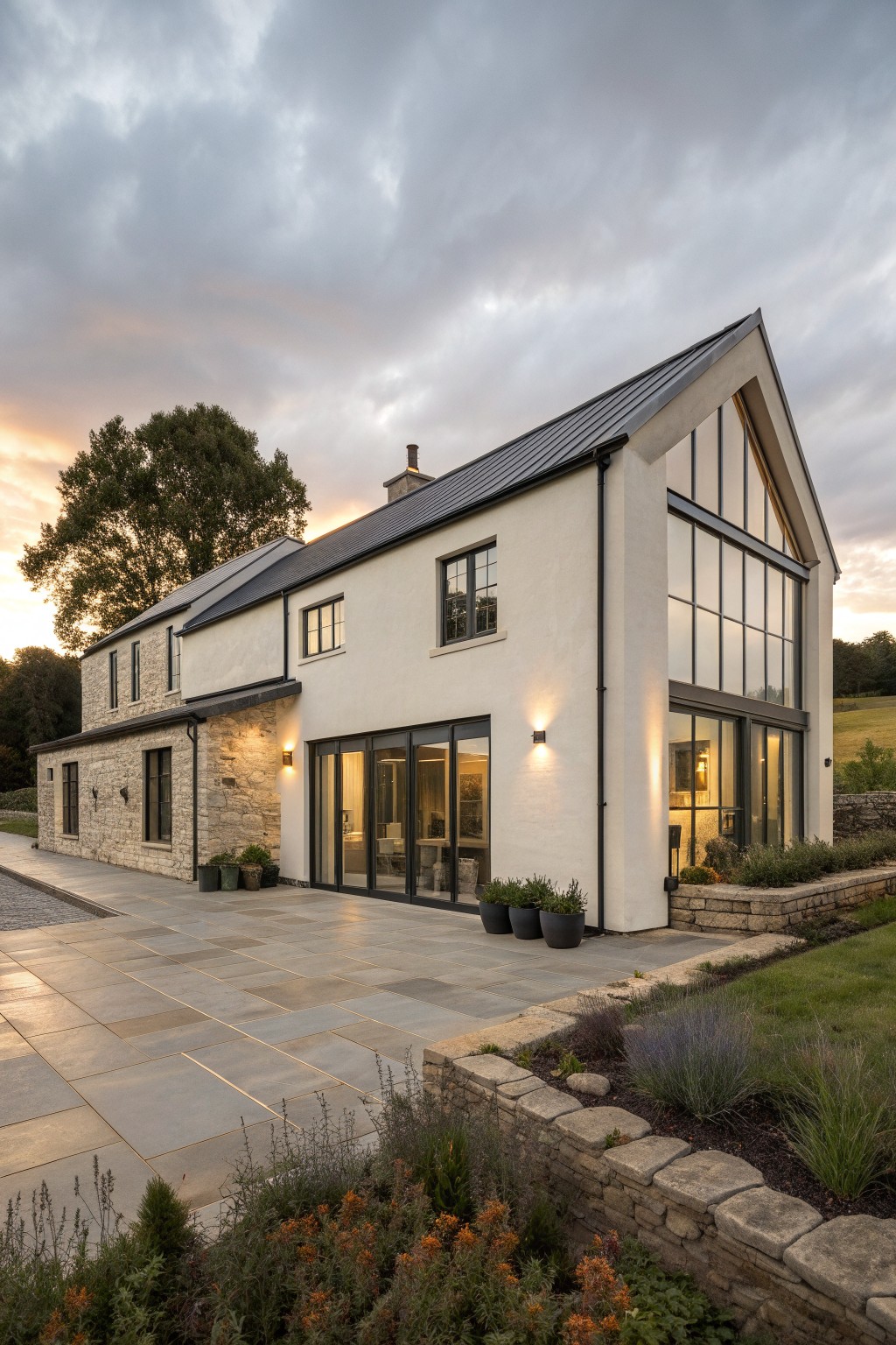 A two-story European farmhouse exterior with white render, stone sections, dark metal roof, and large black-framed windows and doors opening onto a stone patio.