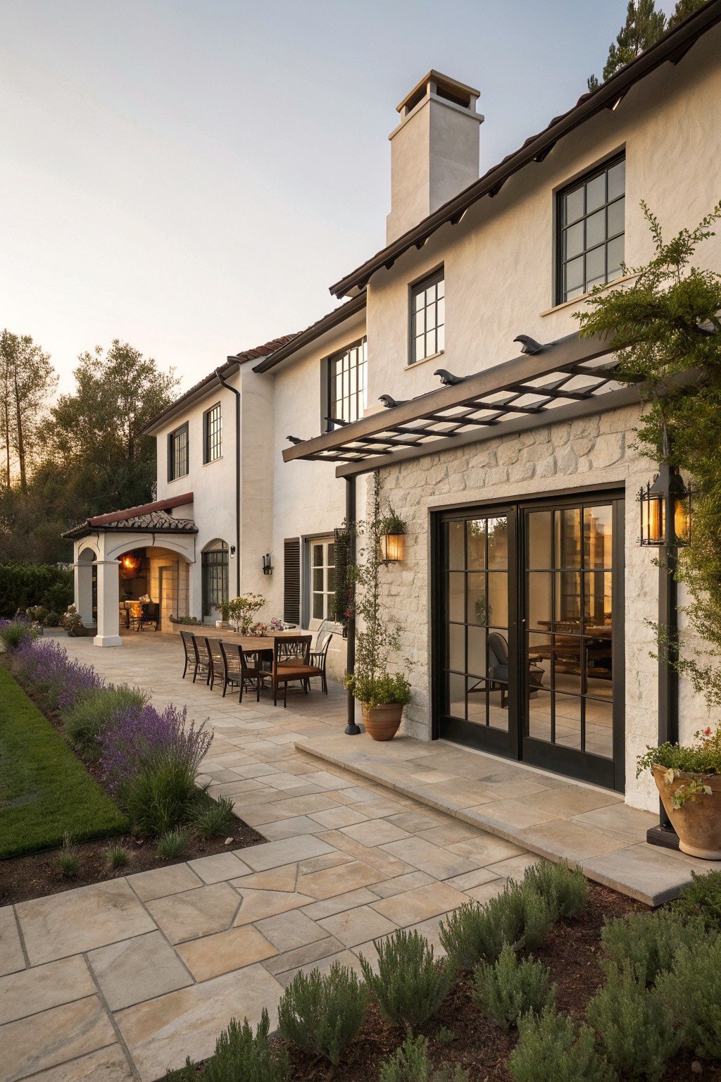 A two-story European farmhouse with white stucco walls, stone accents, dark-framed windows and doors, and a stone patio with outdoor dining furniture.