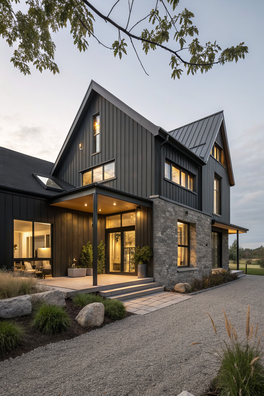 A two-story modern farmhouse with dark vertical siding, a stone base on the right side, a covered front porch, and a gravel driveway.