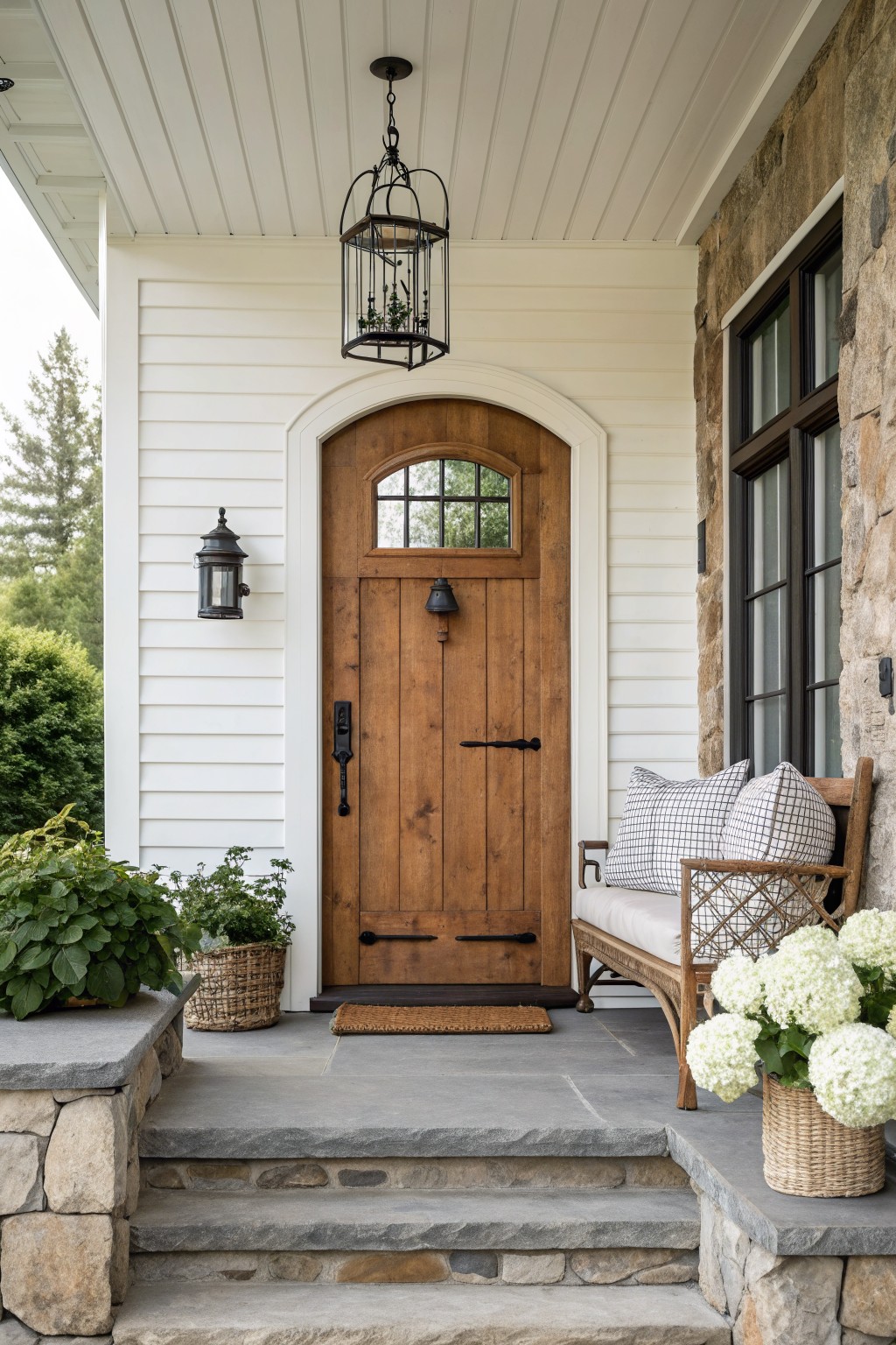 Stone steps to arched wooden door on porch with wicker bench and plants.