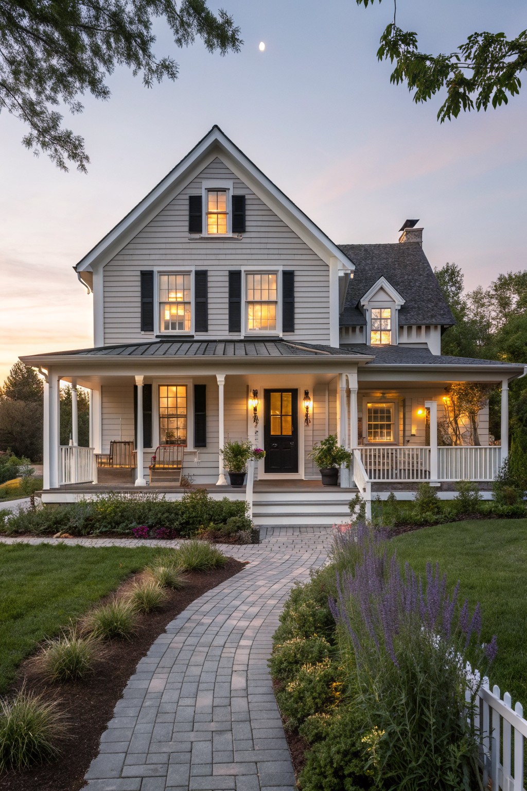 Two-story white house with black shutters, lit windows, porch, and brick path at dusk