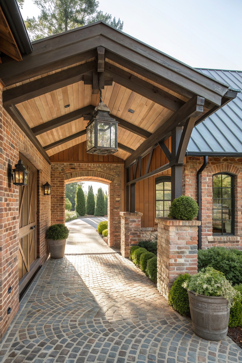 A brick farmhouse entry with dark timber beams supporting a covered walkway over a cobblestone path, featuring a hanging lantern and an arched brick opening in the background.
