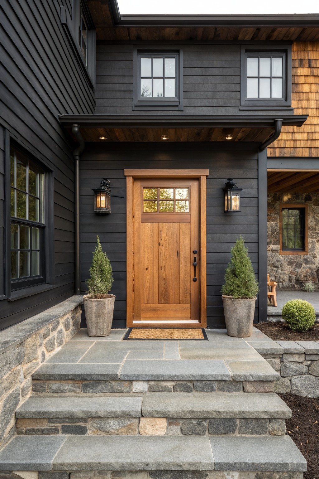 Front view of a dark wood-sided house with a centered cedar door, two wall lanterns, stone steps, and potted shrubs on either side of the entrance.