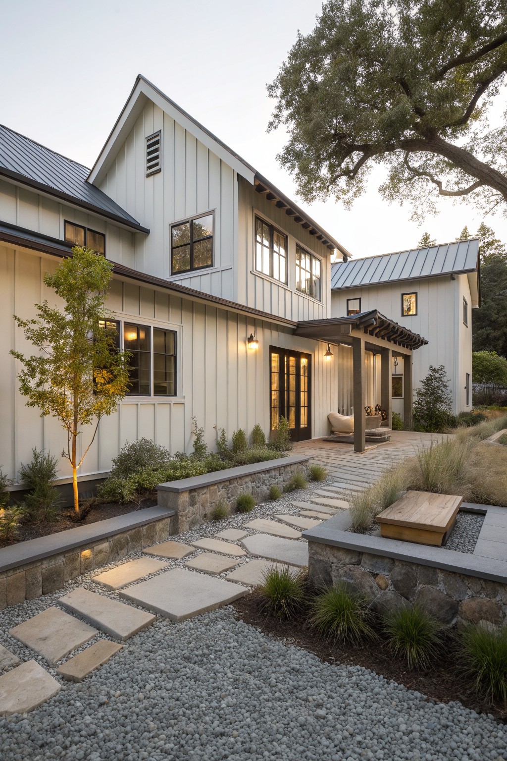 A white farmhouse exterior with metal roof, a gravel path of large stepping stones, stone retaining walls, and a wooden platform in the landscaped foreground.