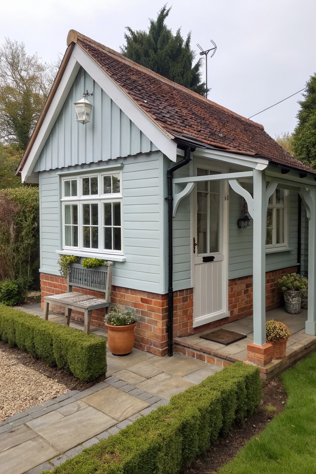 Pale blue wooden shed with tiled roof, white door, porch, and potted plants.