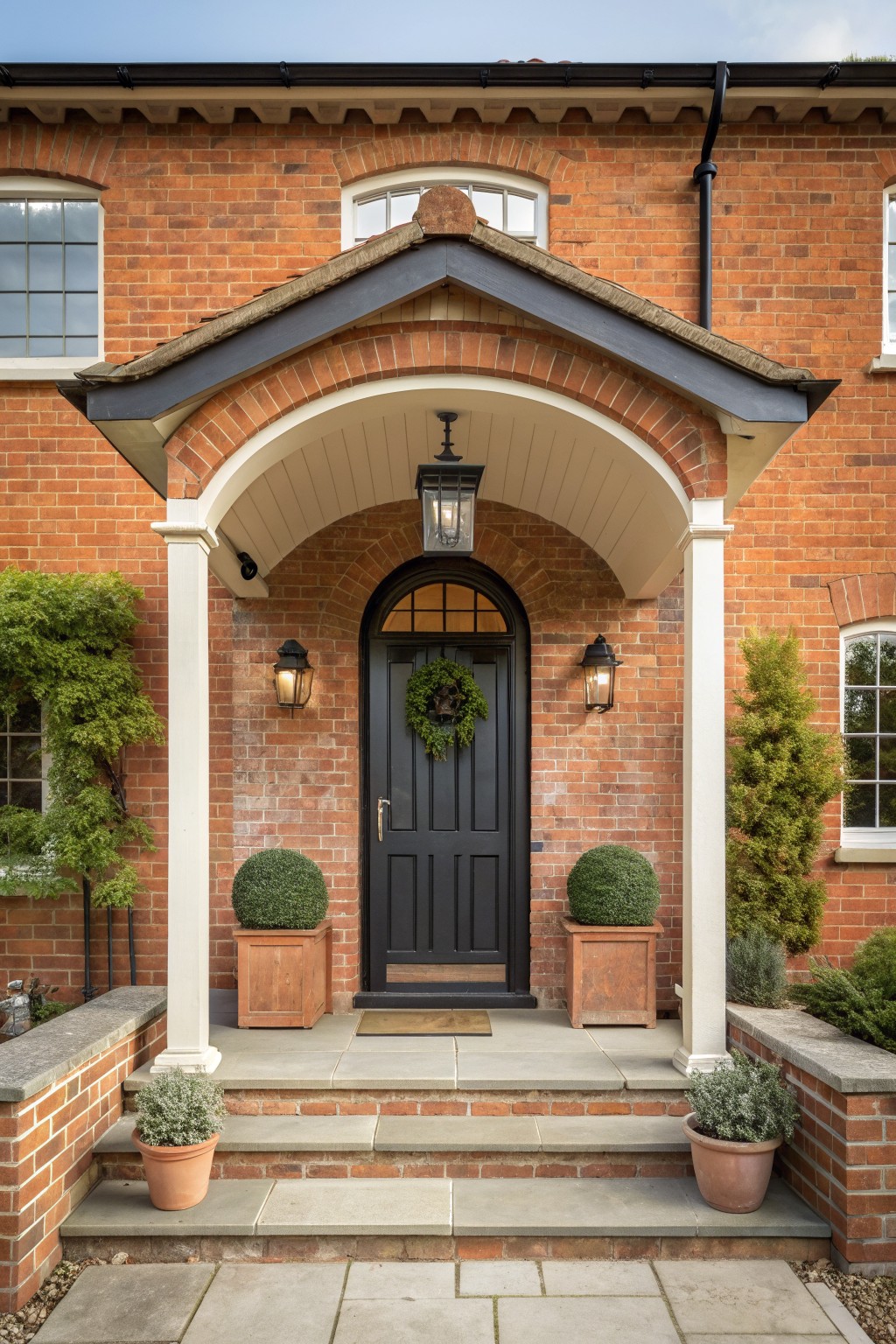 Front view of a red brick house with a dark arched brick entry, black paneled door, and two wooden planters with round shrubs flanking the steps.