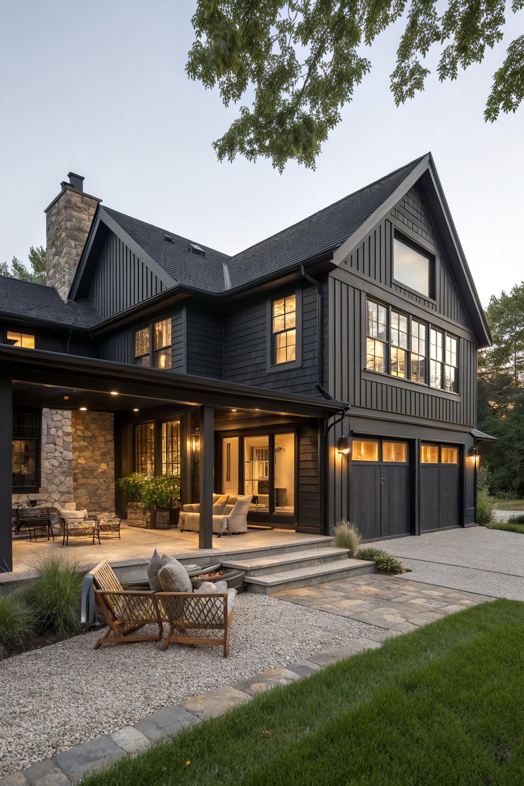 A two-story house with black siding, stone chimney, gabled roof, and a covered patio with seating next to a gravel fire pit area.