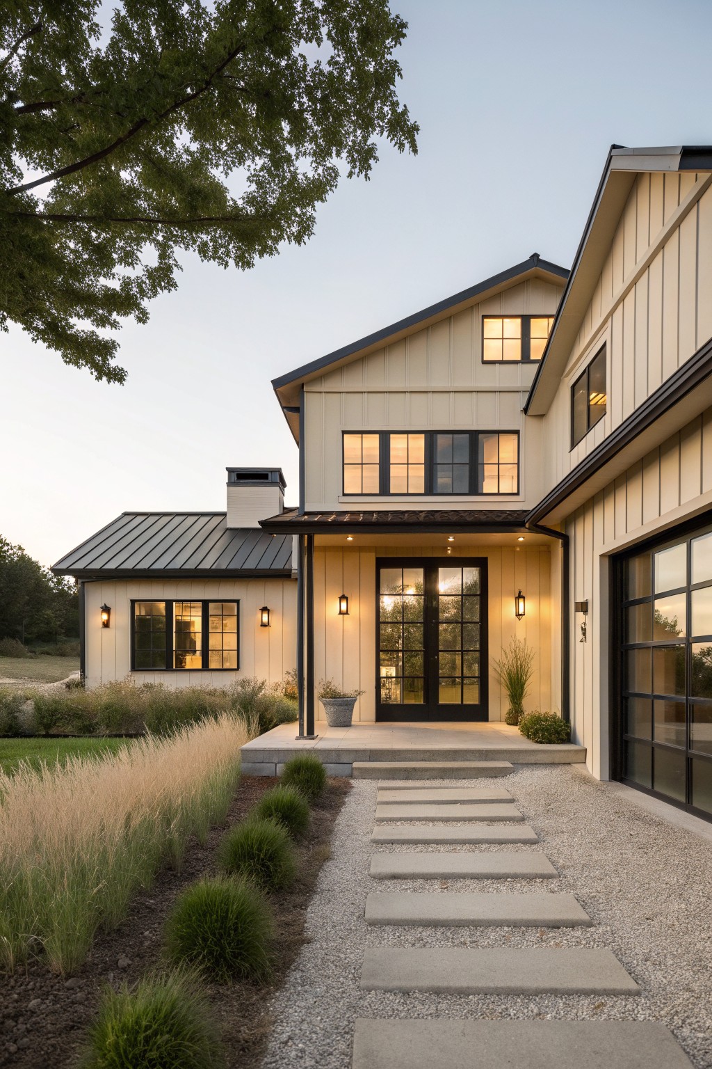 A two-story farmhouse with light beige siding, black-framed windows and doors, a dark metal roof on the left wing, and a gravel path with stepping stones leading to the front entrance.