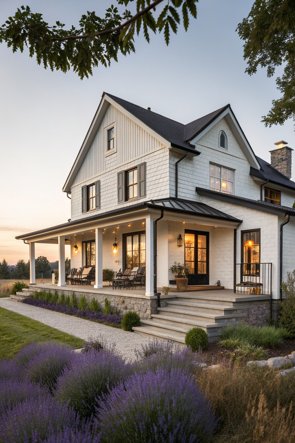 White two-story farmhouse with porch lights, stone steps, and blooming purple lavender at dusk.