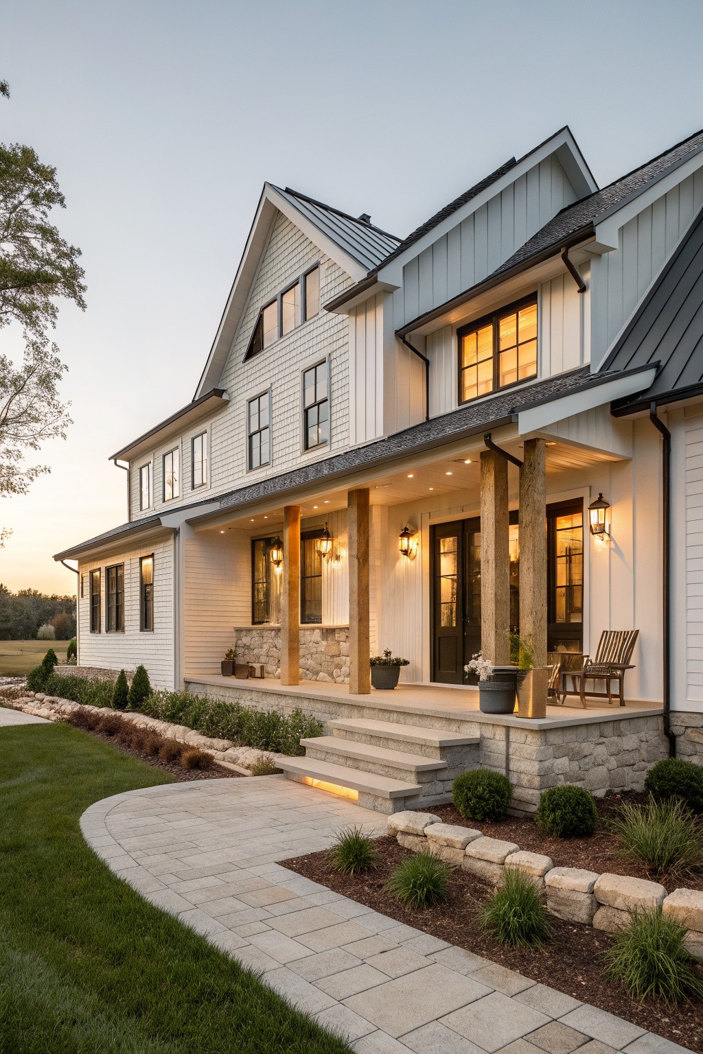 Large white modern farmhouse with stone porch, wooden columns, and lit windows at dusk.