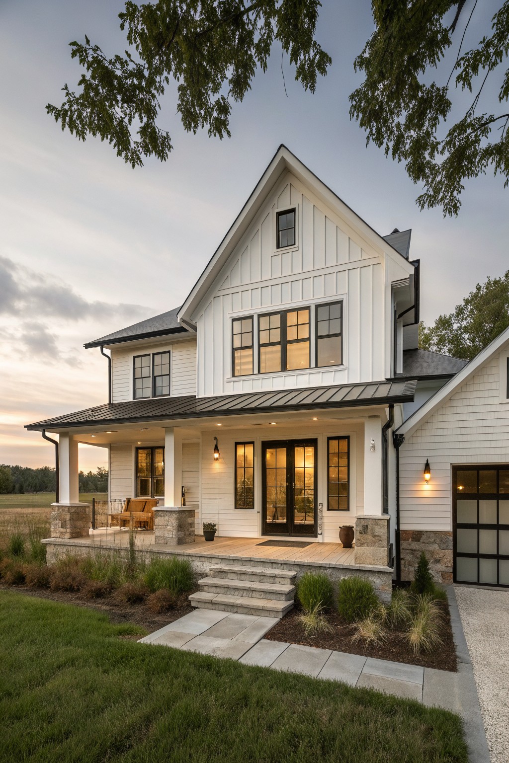 A white two-story farmhouse exterior with board and batten siding, a dark metal roof, stone bases on the porch columns, and a covered front porch with steps.