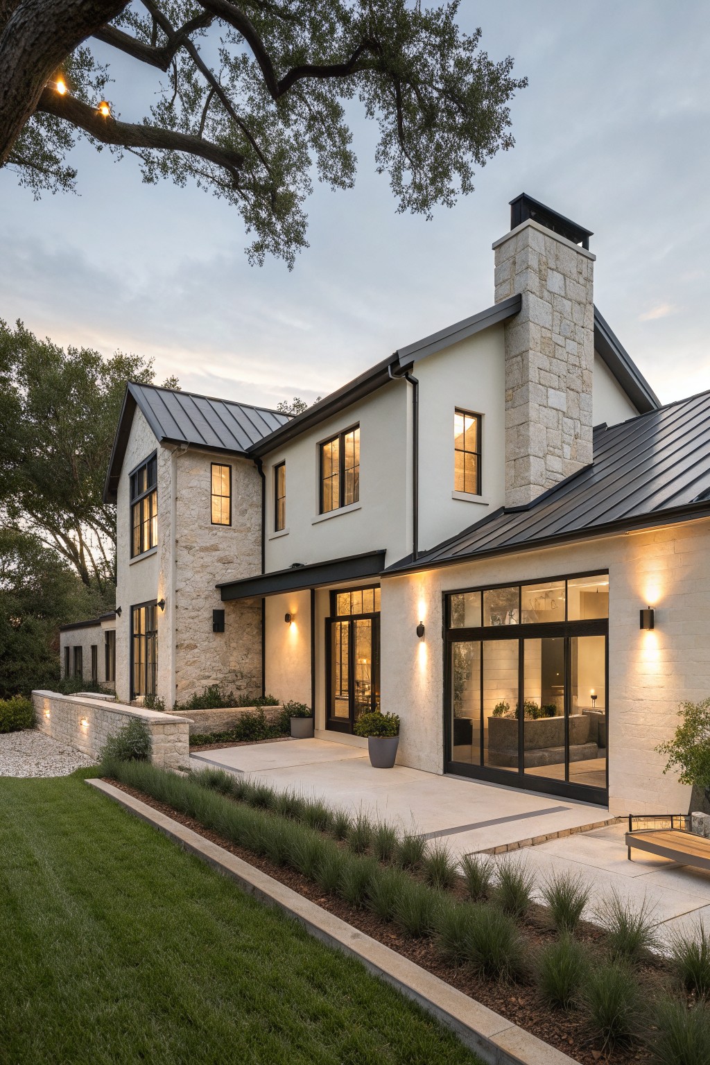 A two-story modern European farmhouse exterior with white walls, stone accents, a stone chimney, dark metal roof, and large black-framed windows and doors opening to a patio and landscaped yard.
