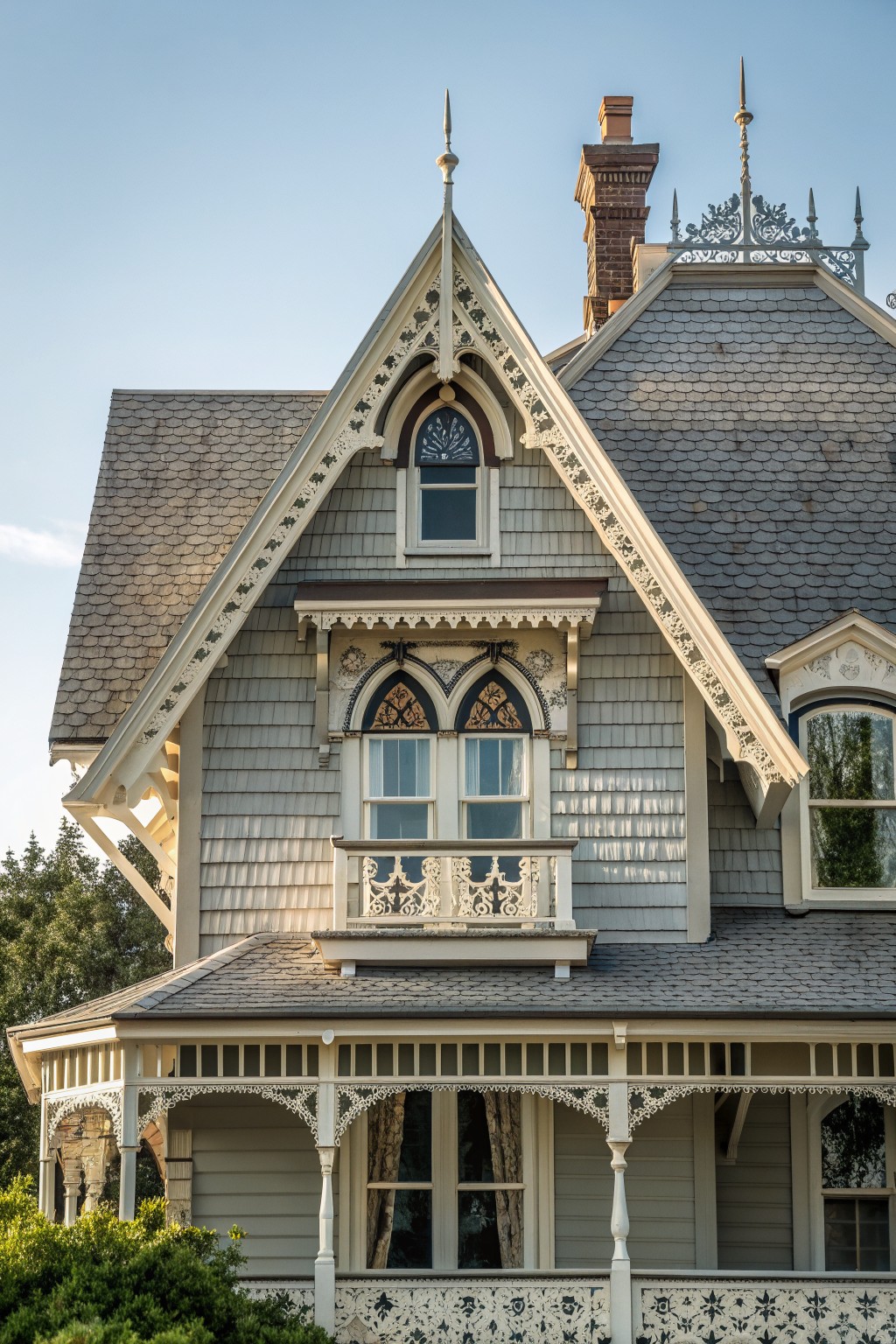 Close view of a gray Victorian farmhouse with a tall gable featuring white decorative trim, an arched window, and a small balcony below.