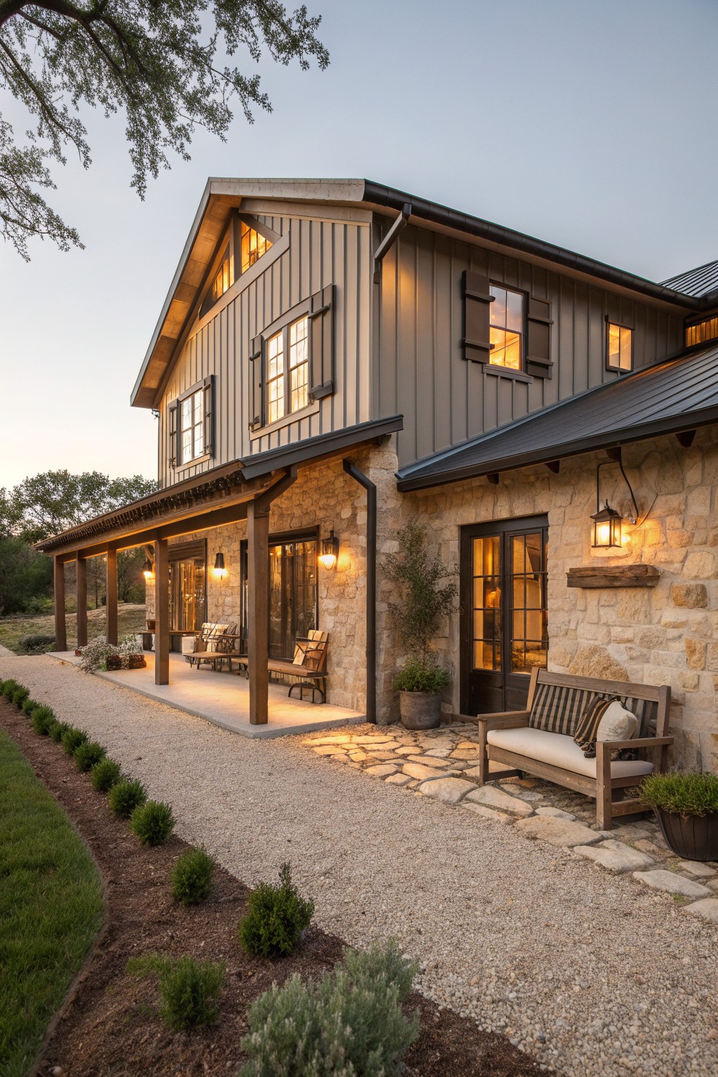 Modern gray farmhouse with stone walls, wooden porch, bench, and glowing windows at dusk.