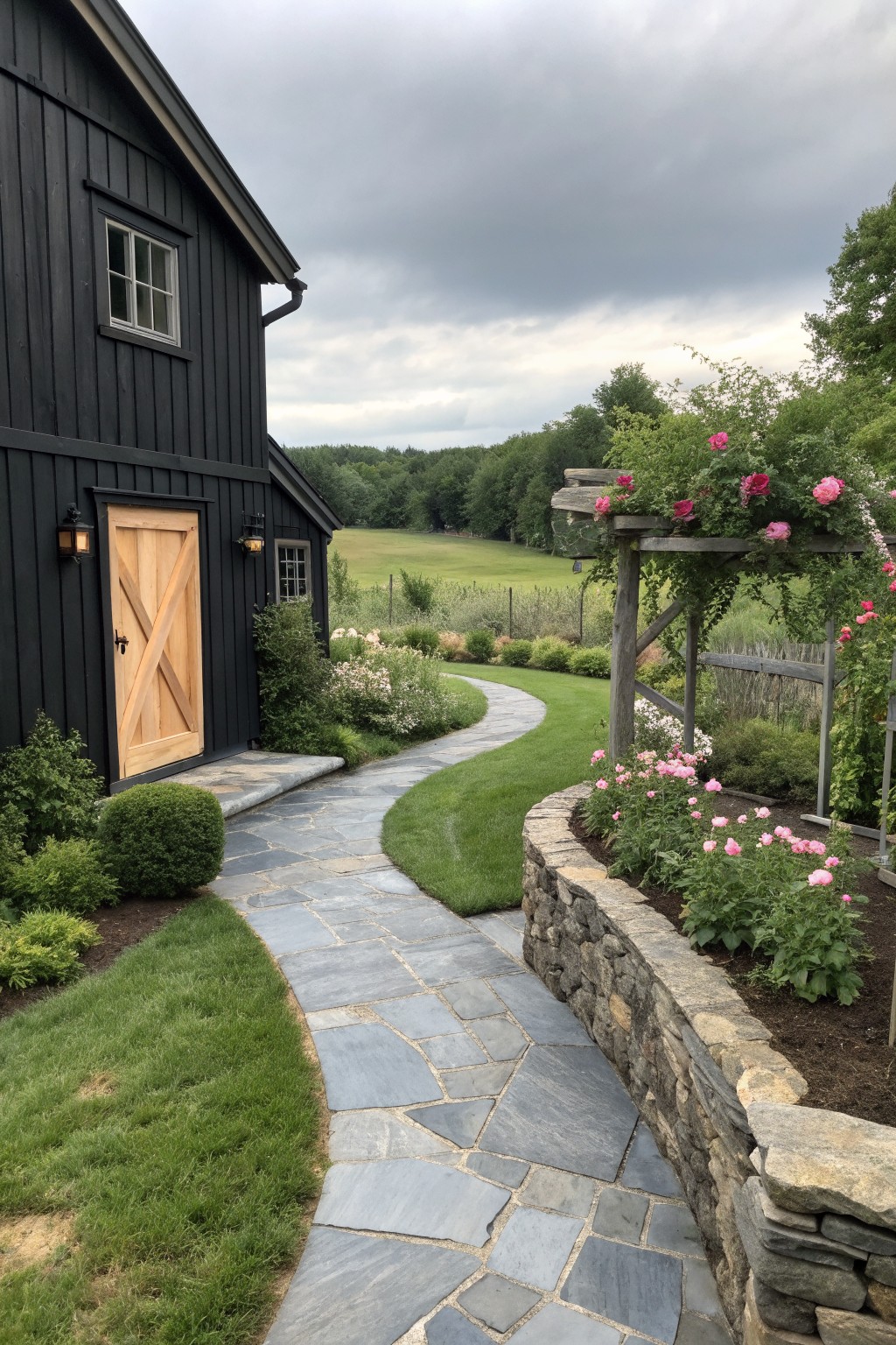 A black vertical siding farmhouse with a light wood door, stone pathway, and garden with pink roses on a wooden pergola.