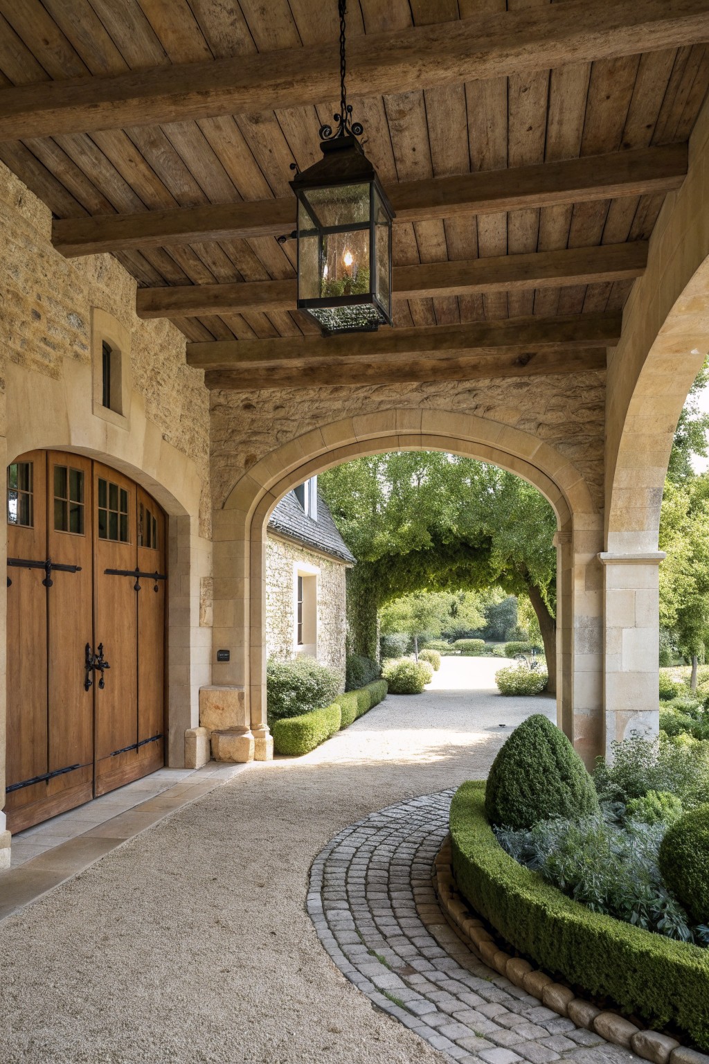 A stone archway covers an entry with wooden double doors on the left, a hanging lantern overhead, and a gravel path that curves past greenery into the garden.