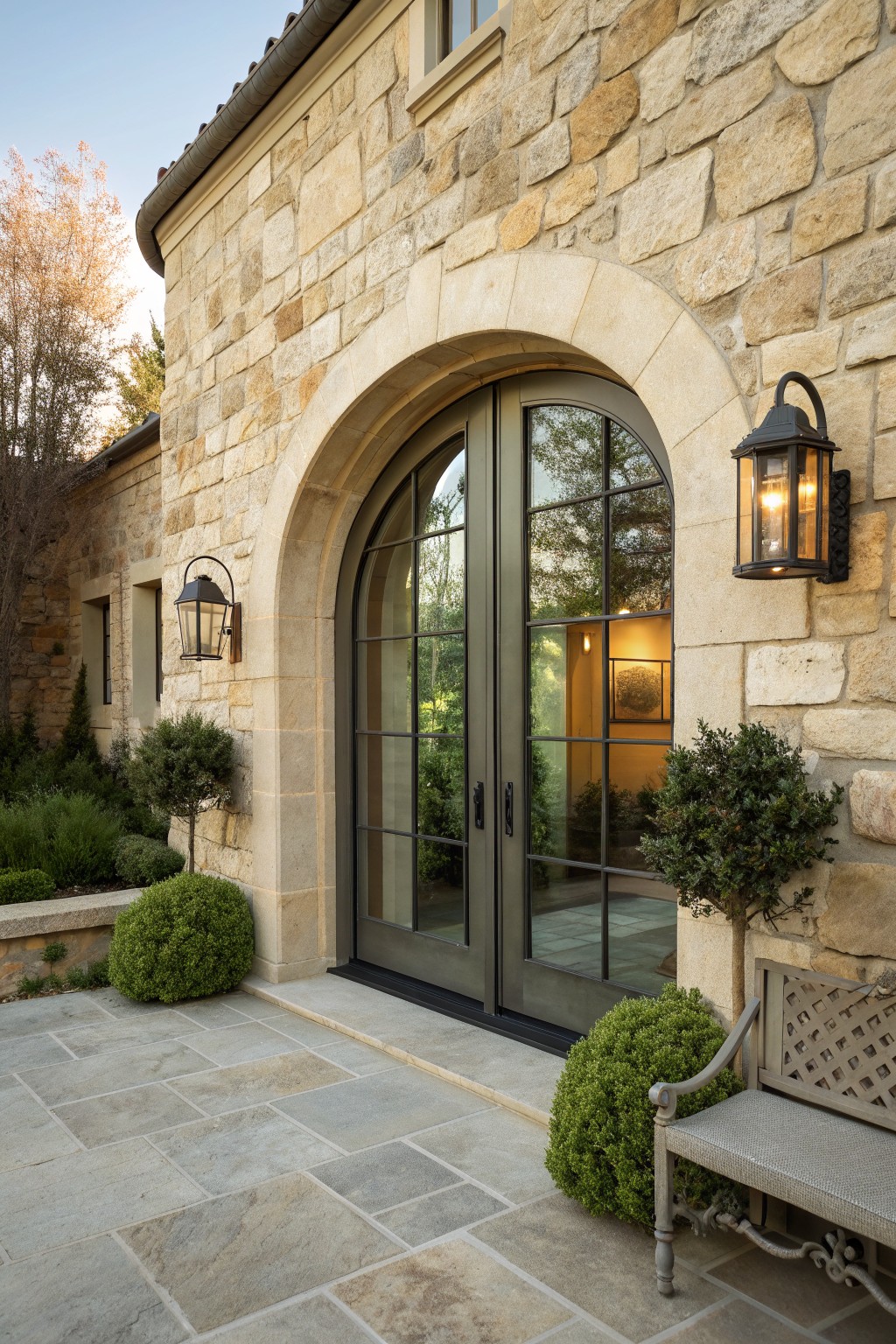 A stone house exterior featuring a large arched entryway with double glass doors, wall lanterns, and a stone bench nearby.