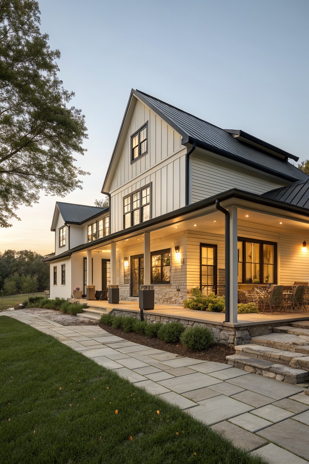 A two-story white farmhouse with board and batten siding, black metal roof, covered porch, and stone accents viewed from the front lawn at dusk.