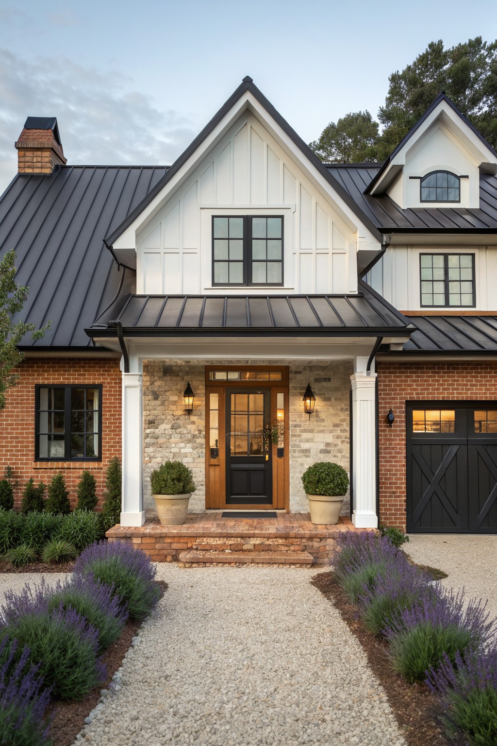 A front view of a two-story farmhouse with a black metal roof, white gables, brick walls, stone entry, and a gravel path lined with lavender.