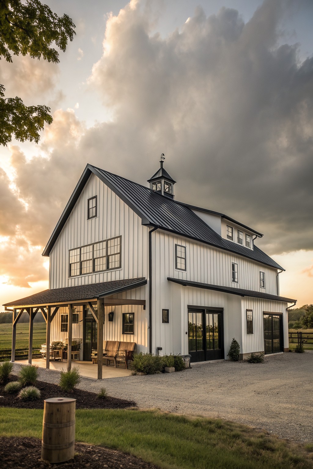 A white two-story farmhouse with black metal roofing, a cupola, and a side porch, viewed from the gravel driveway at dusk.