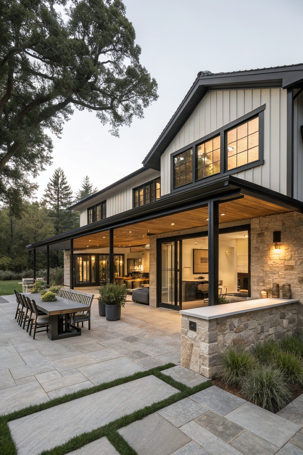 A modern farmhouse exterior with black metal roof, white vertical siding, black window trim, and a large covered patio featuring a long outdoor dining table on stone pavers.