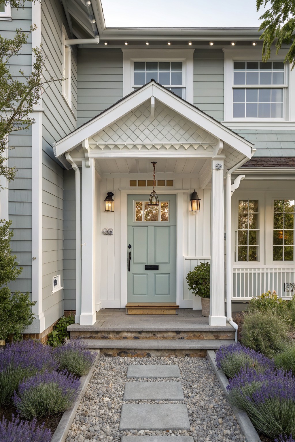 Front view of a light gray house with white trim, a gabled porch supported by columns, a sage green door, and a gravel path with concrete stepping stones leading to the steps.