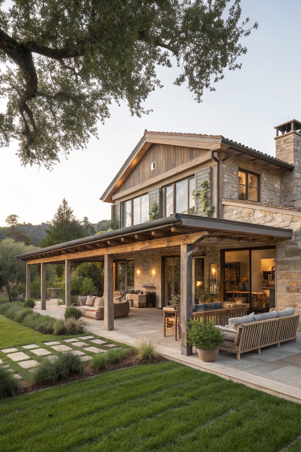 A rustic modern farmhouse with stone walls and wood siding features a wide covered wooden porch with multiple seating areas overlooking a lawn and garden.