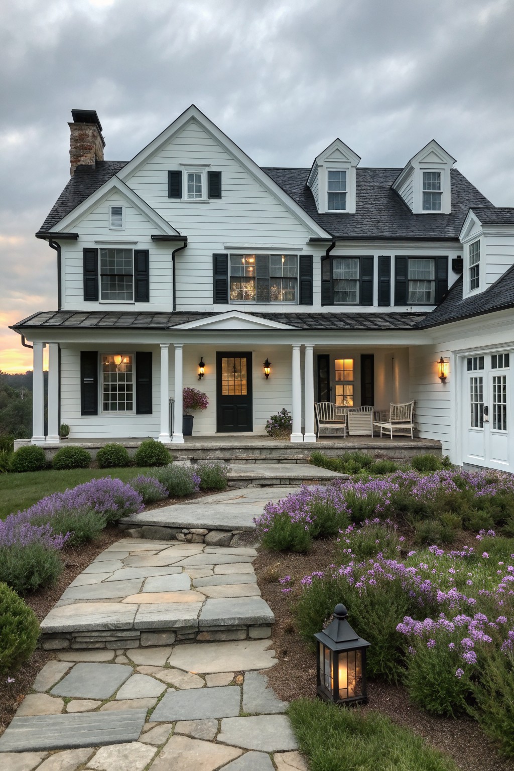 A large white two-story farmhouse with black shutters, a central black front door, a covered porch with white columns, and a stone pathway leading through purple flowering shrubs.