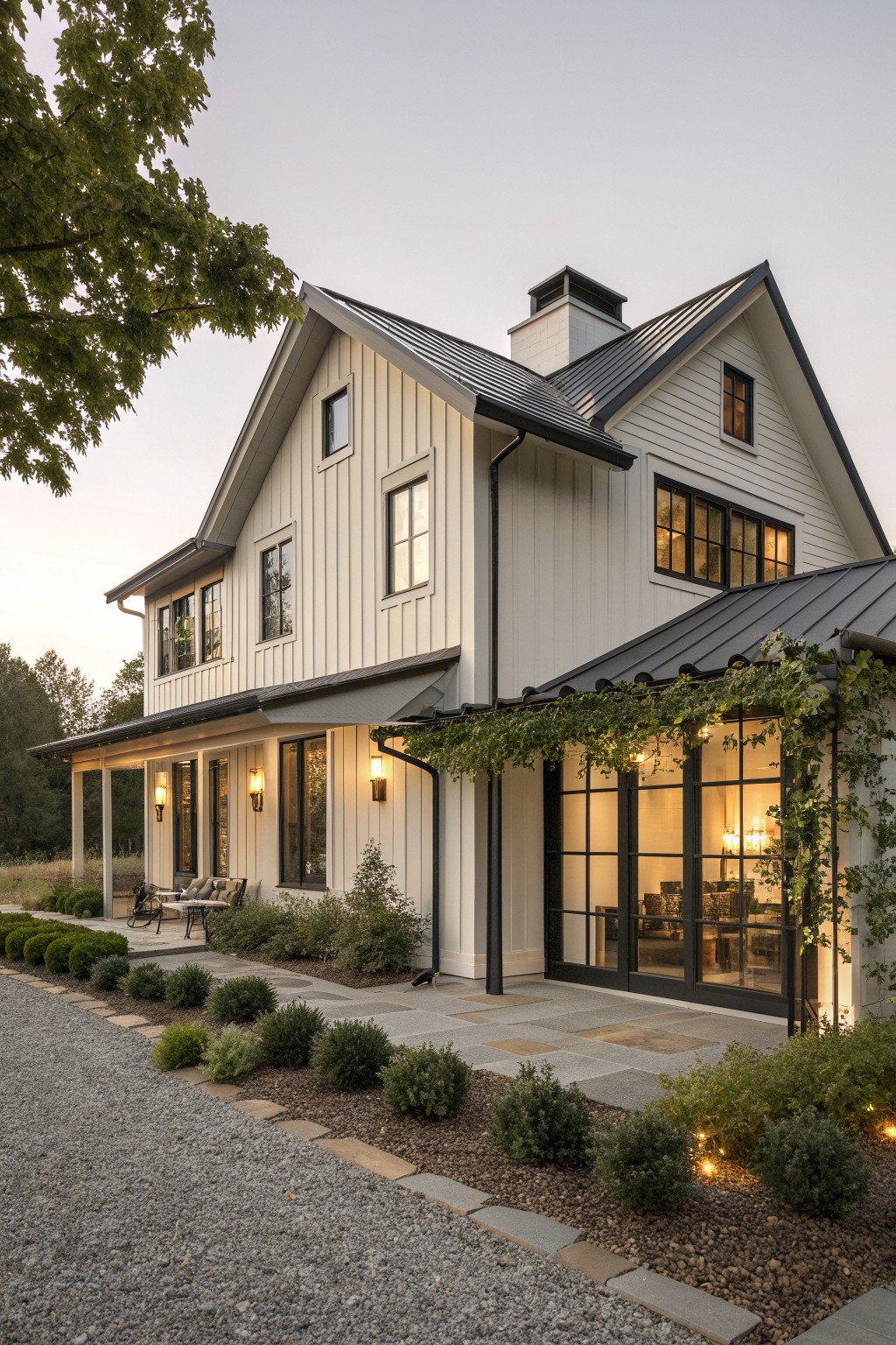 A two-story farmhouse exterior with light vertical siding, dark metal roofing, black-framed windows, and a stone patio area with surrounding landscaping.