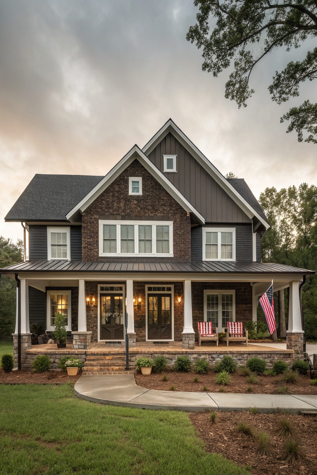 A two-story farmhouse exterior with dark gray siding, brick sections, white trim, a covered porch with white columns, and two red and white striped chairs on the porch.