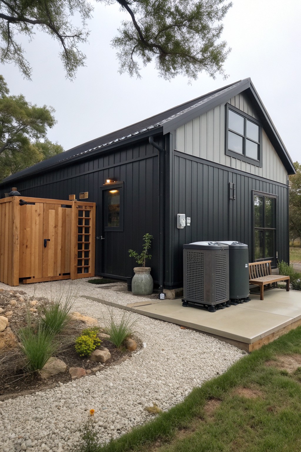 A small barndominium exterior with black vertical siding on the lower walls, light gray siding above, a dark metal roof, and a gravel path beside a wooden enclosure.