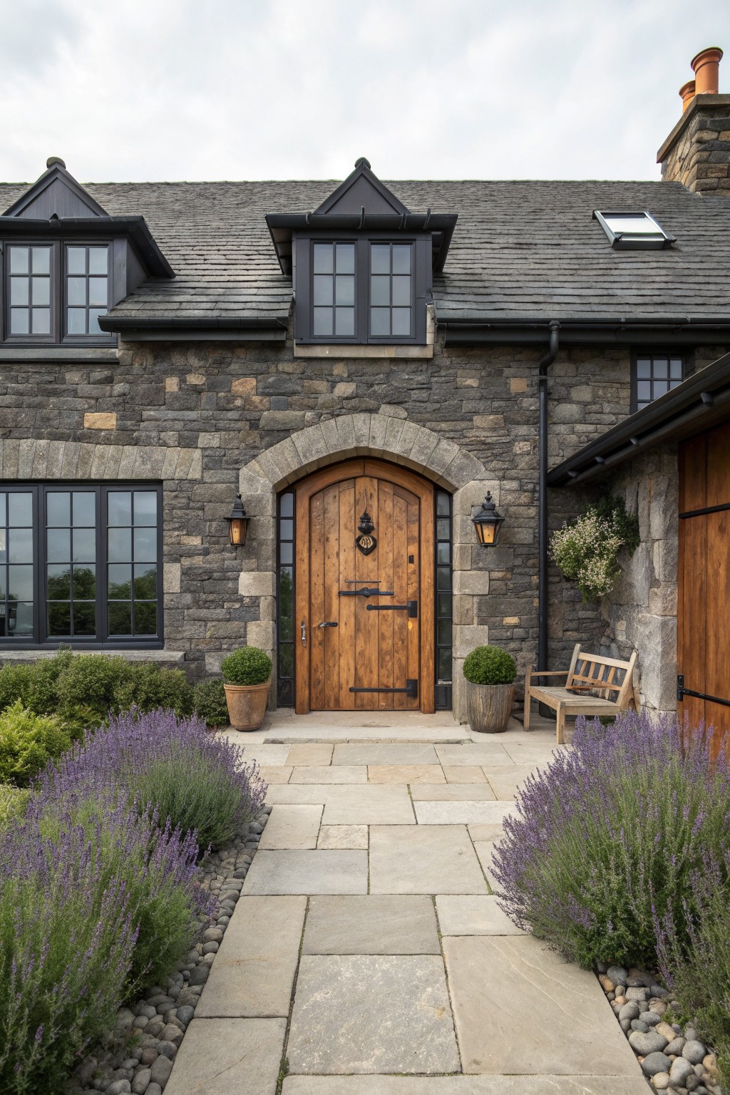 A stone house exterior featuring black-framed windows, an arched wooden door, and purple flowering plants beside a stone pathway.