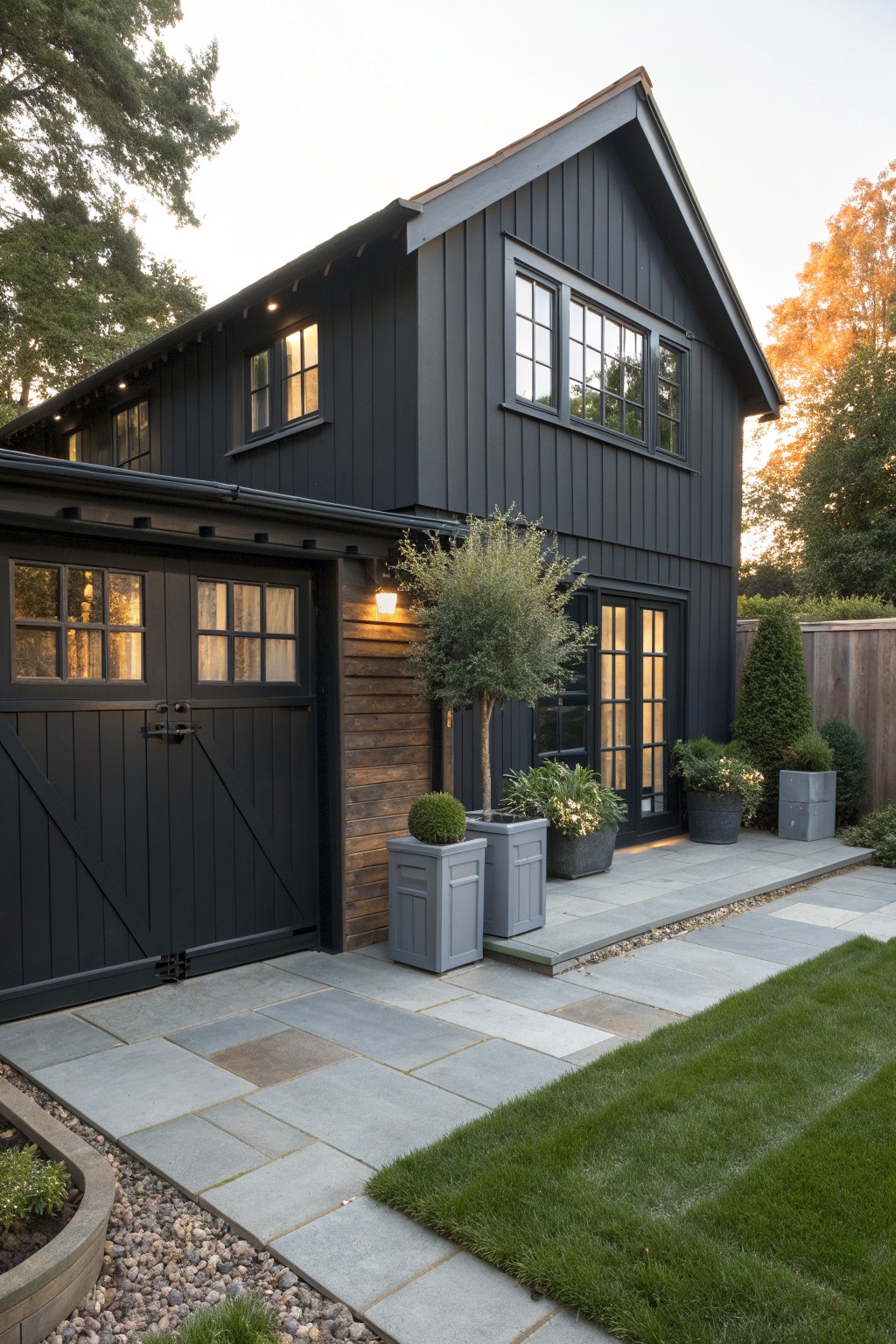 A two-story house with black vertical siding, black garage doors, black-framed windows, a stone patio, and several potted plants in front.