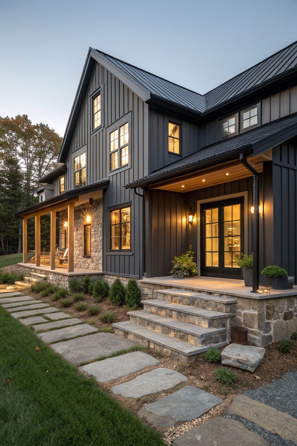 A modern two-story house with dark vertical siding, a stone foundation, wood porch posts, a metal roof, and stone steps leading to a lit entry at dusk.