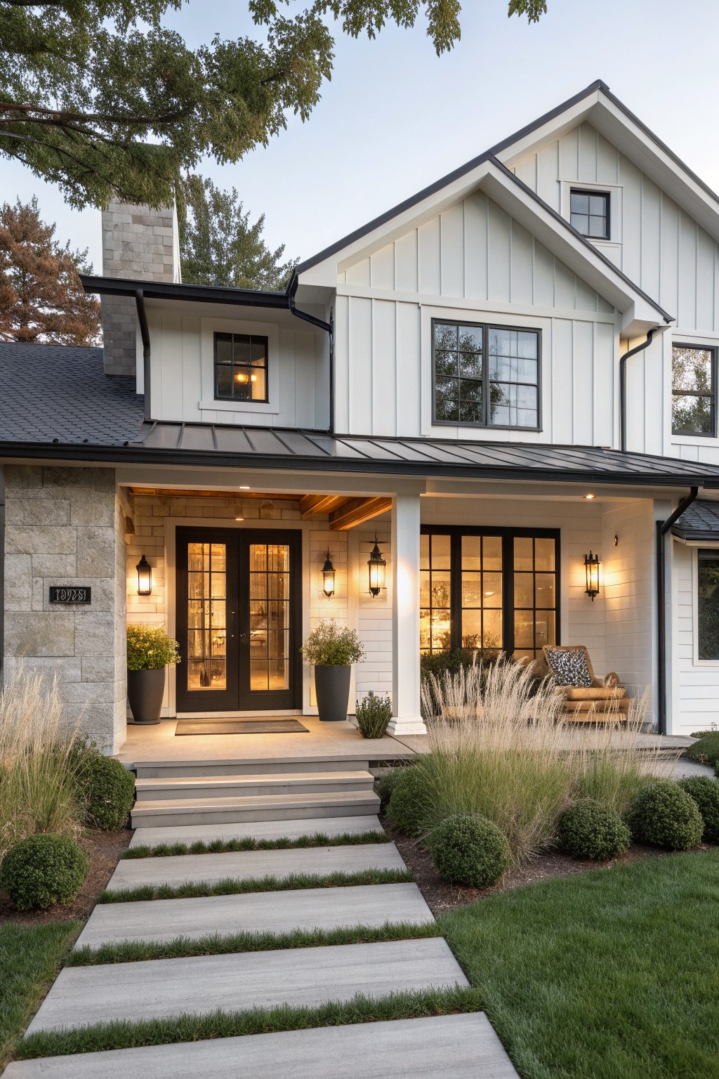 Modern white farmhouse with black trim, lit porch, concrete steps, and landscaping at dusk.
