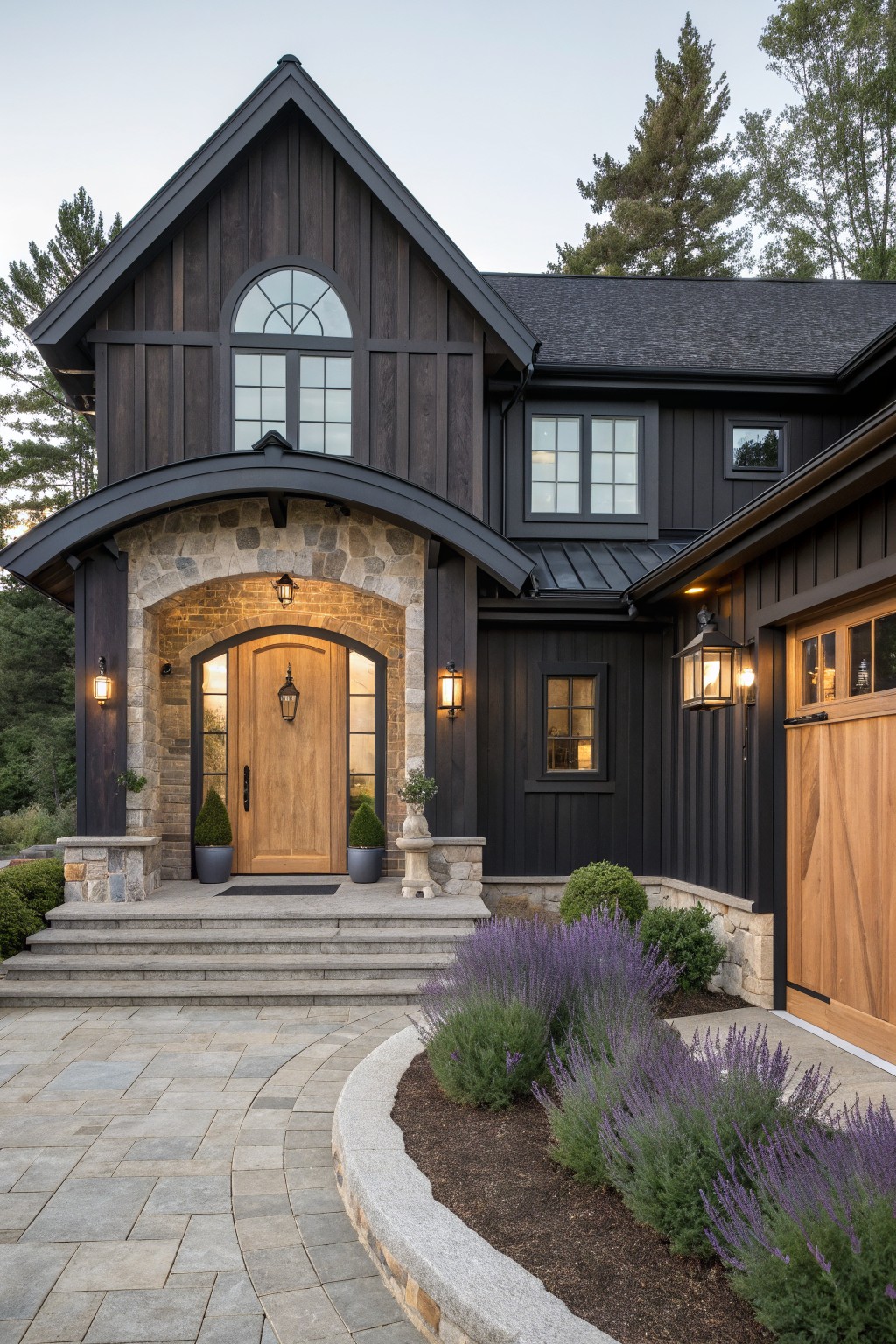 Front view of a dark board and batten house with a large arched stone entry surrounding a wooden door and steps leading up from a paved driveway.