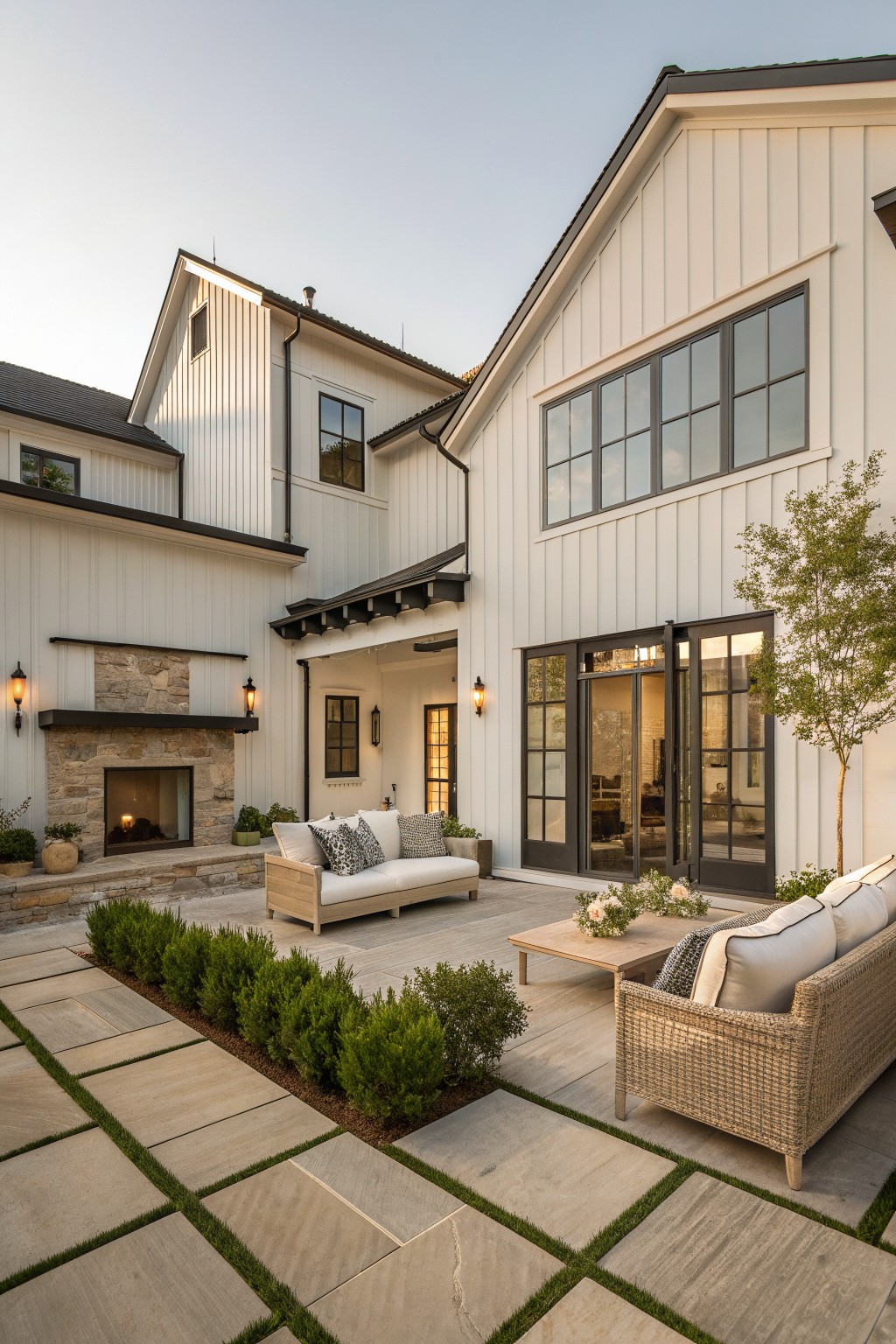 White board and batten farmhouse exterior with dark trim, multiple gables, stone outdoor fireplace, and paved patio with seating.