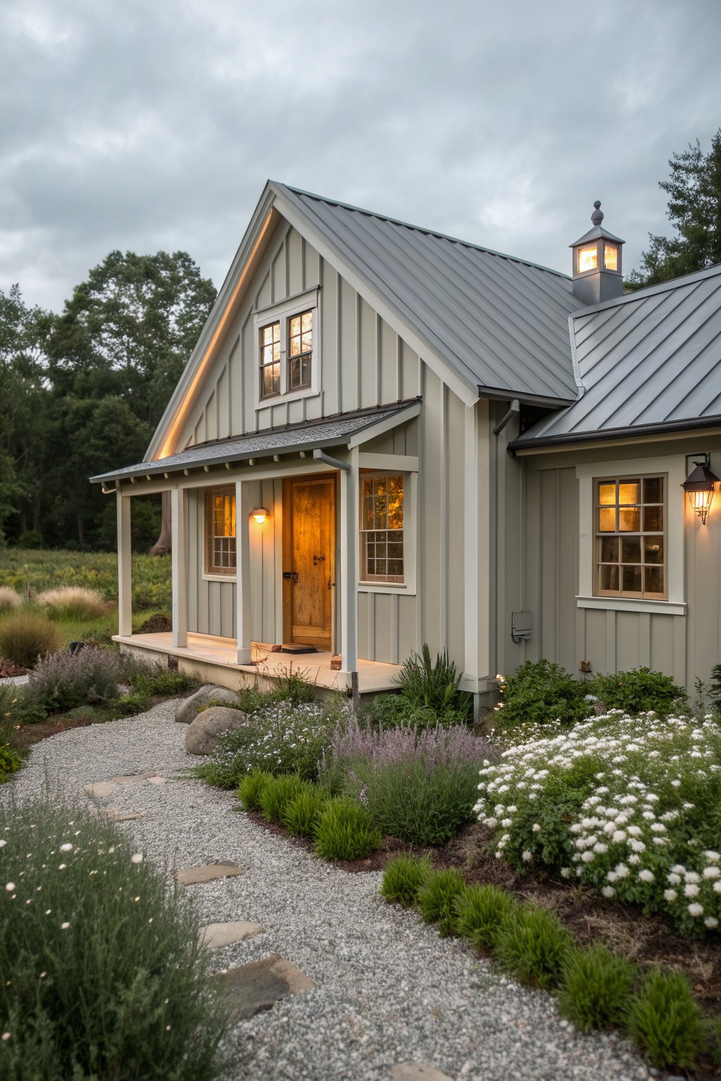 Gray cottage with metal roof, glowing windows, wooden door, and flower-lined gravel path at dusk.
