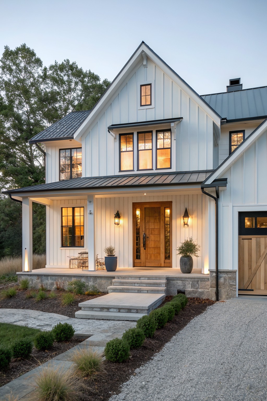 A white board and batten farmhouse with a large front gable, wooden door, stone porch base, and black metal roof accents at dusk.