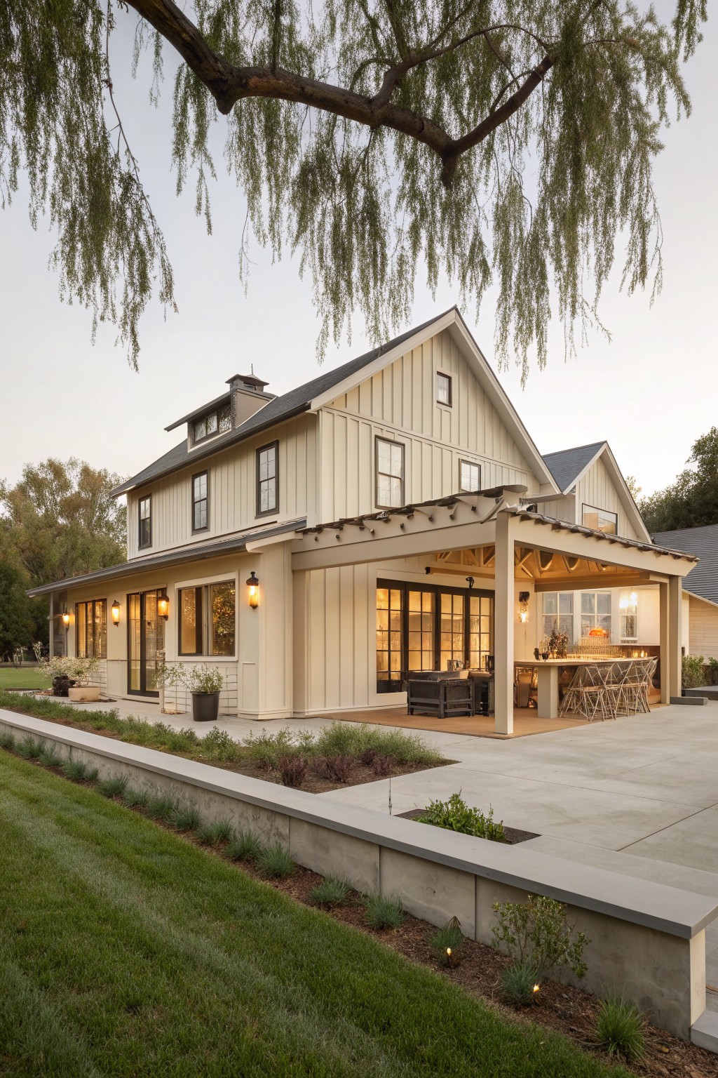 A two-story farmhouse with light board and batten siding, gabled roofs, and a covered patio with an outdoor dining table.