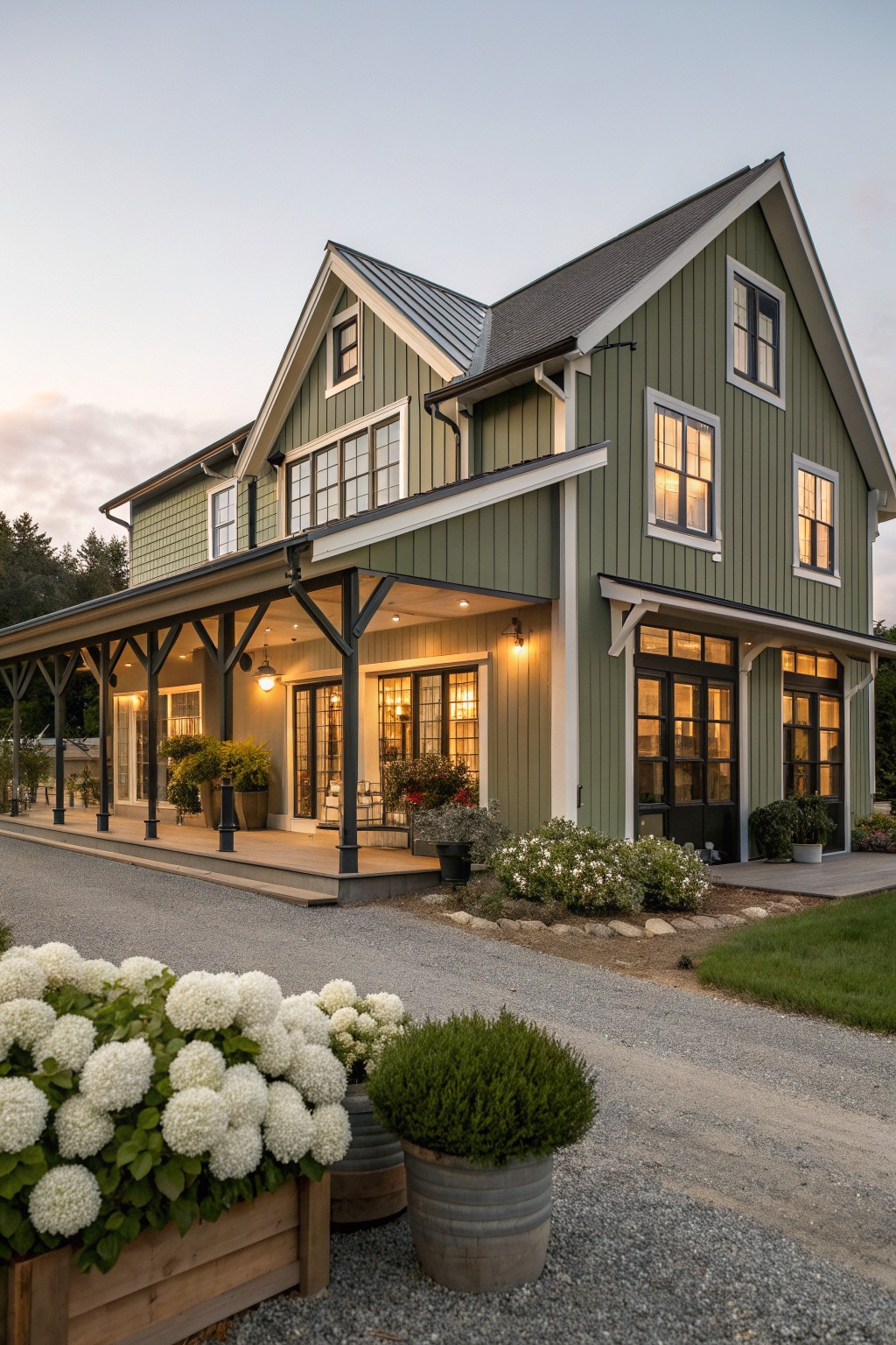 Large green house with covered porch, glowing windows, and potted white flowers on gravel.