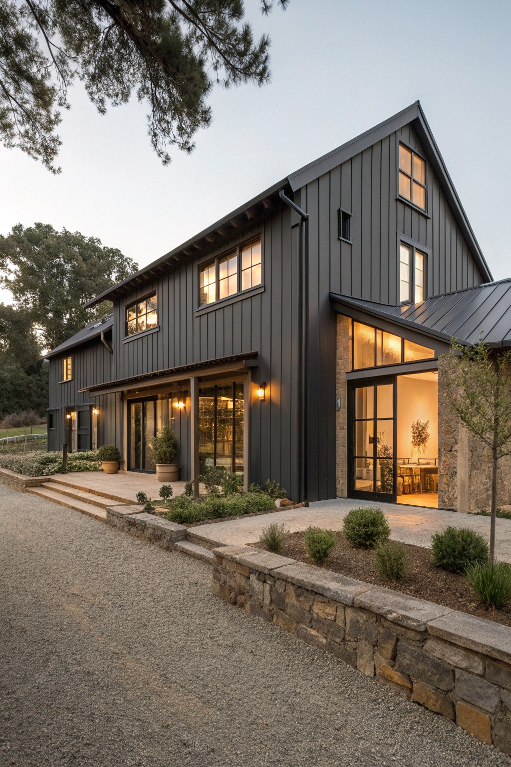 A dark gray modern farmhouse exterior with vertical siding, stone accents, large black-framed windows, and a gravel driveway bordered by low stone walls.